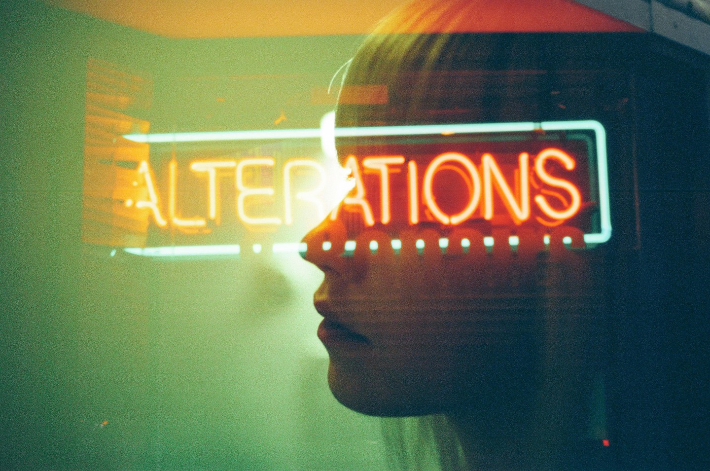 A woman's profile is visible through a window with a neon "ALTERATIONS" sign reflected on it.