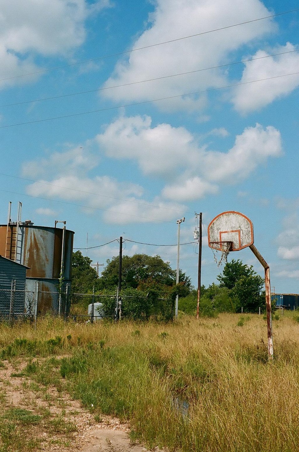 An abandoned basketball hoop stands in a field of tall grass next to industrial structures under a cloudy sky.