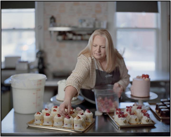 A woman decorates small cakes with fruit and flowers in a bakery kitchen.