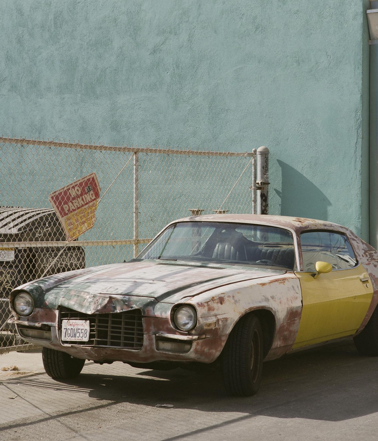A rusty, multi-colored car is parked in front of a chain-link fence and a teal wall with a "no parking" sign.