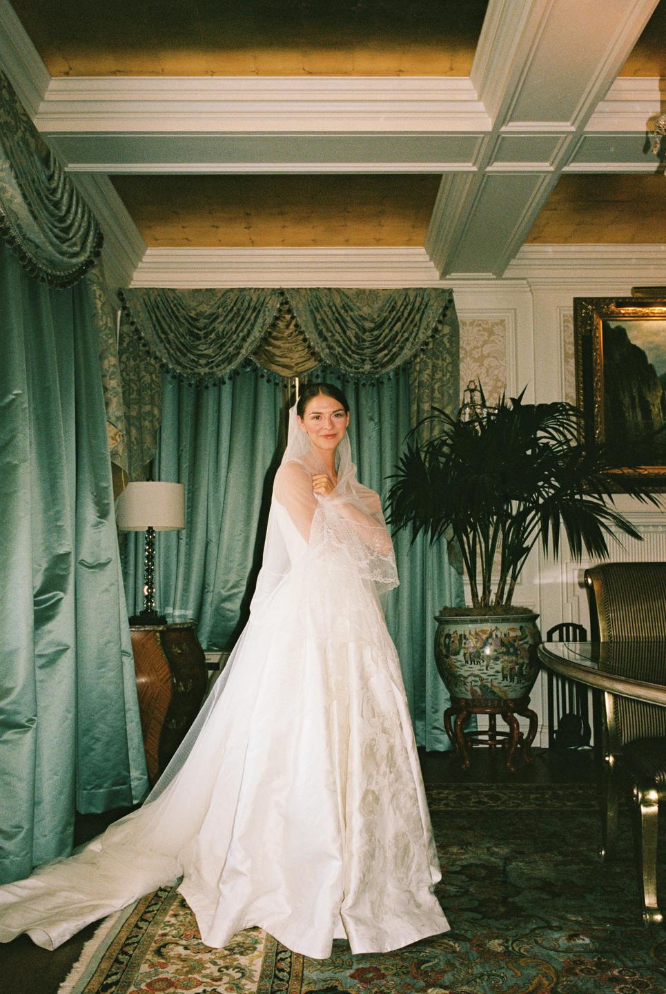 A woman in a wedding dress and veil stands in a room with ornate curtains and a patterned rug.