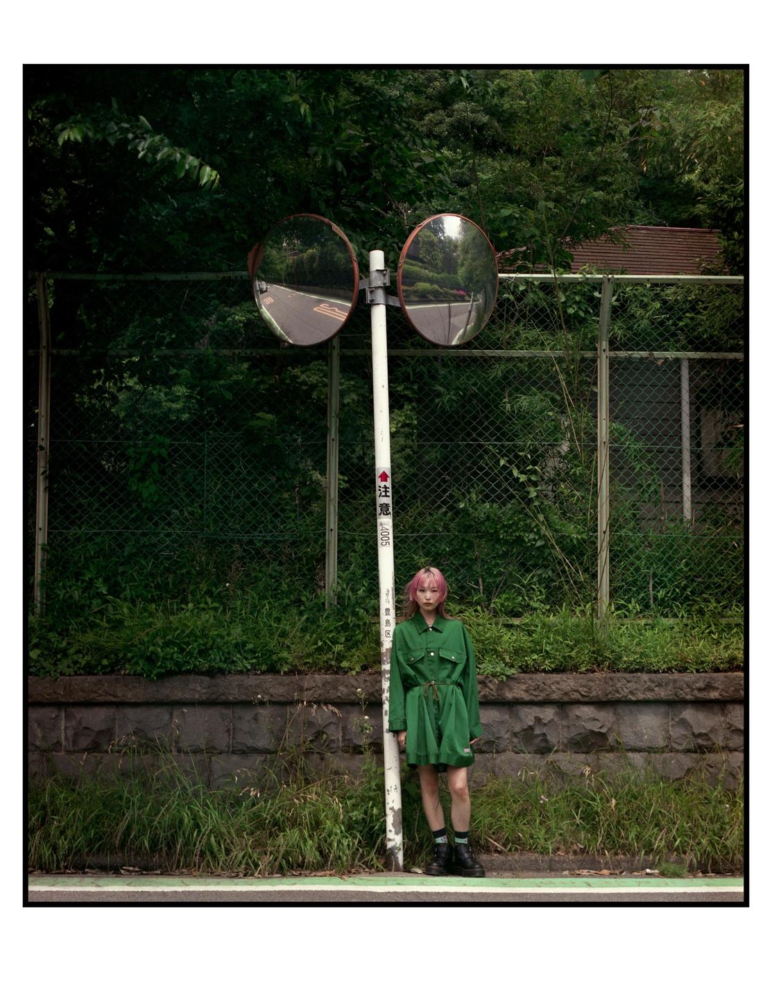 A woman with pink hair stands next to a pole with two convex mirrors on a street.