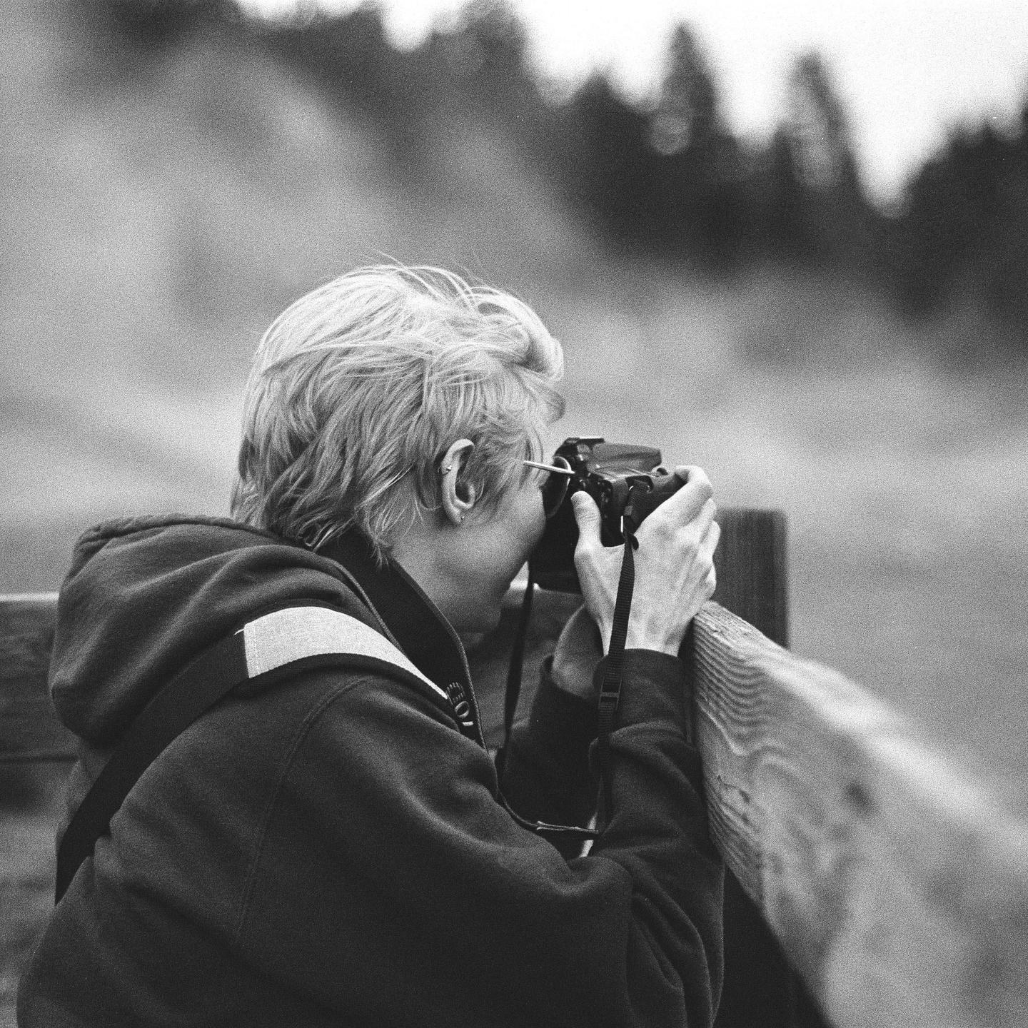 A person with short hair holds a camera to their eye, resting their arms on a wooden fence.