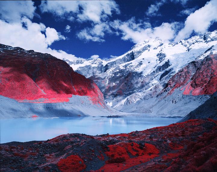 A lake is surrounded by mountains with red foliage and snow-capped peaks under a blue sky with clouds.