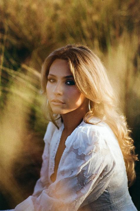 A woman with long blonde hair and a white shirt looks at the viewer in a sunlit field.
