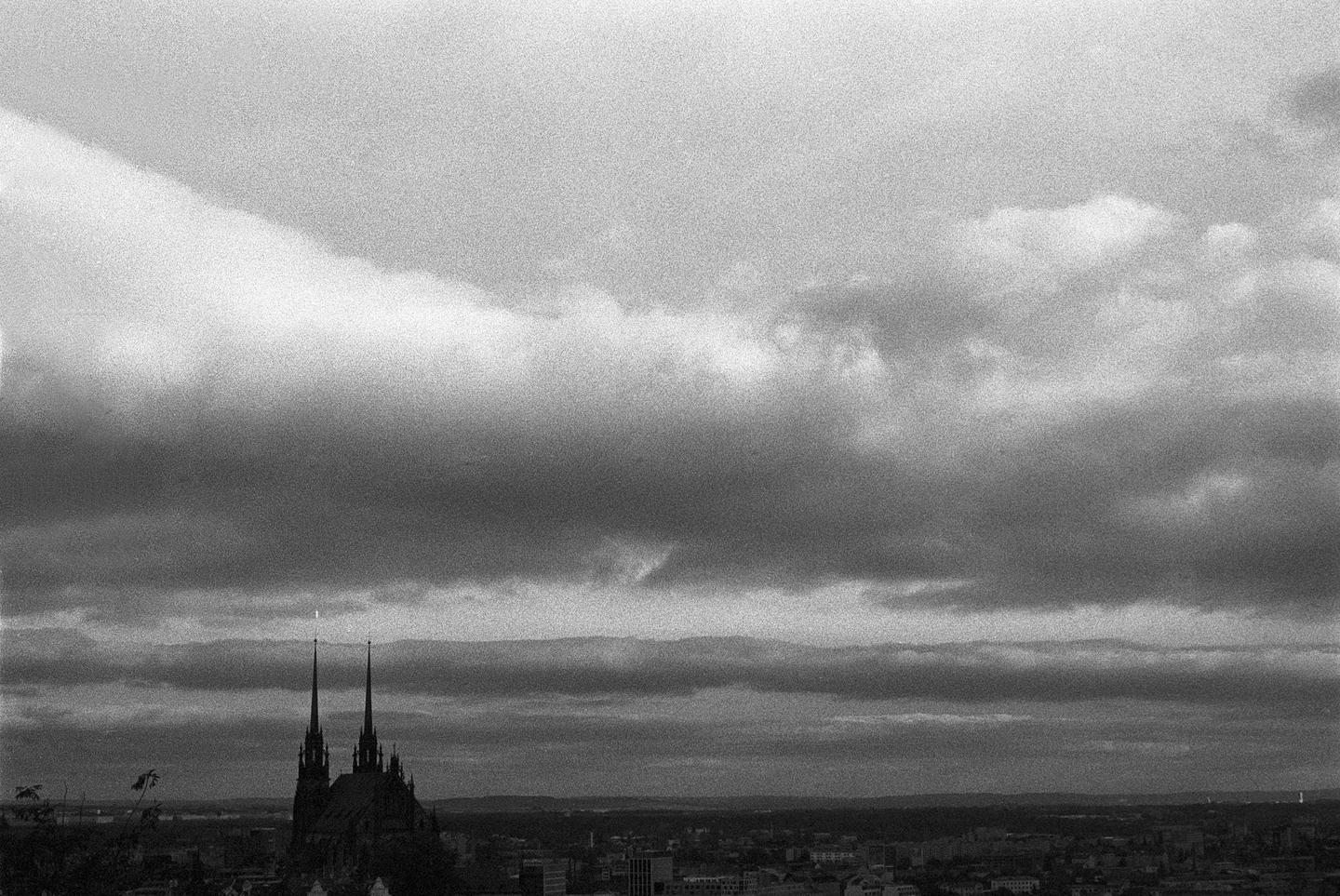 A black and white cityscape shows a cathedral with two spires rising above the buildings under a cloudy sky.