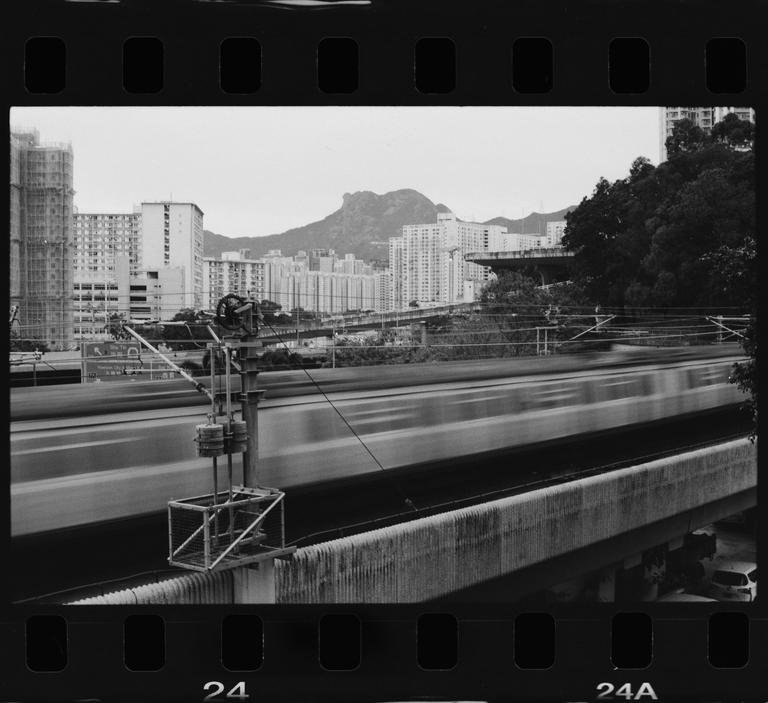 A blurred train moves past a concrete barrier with buildings and a mountain in the background.
