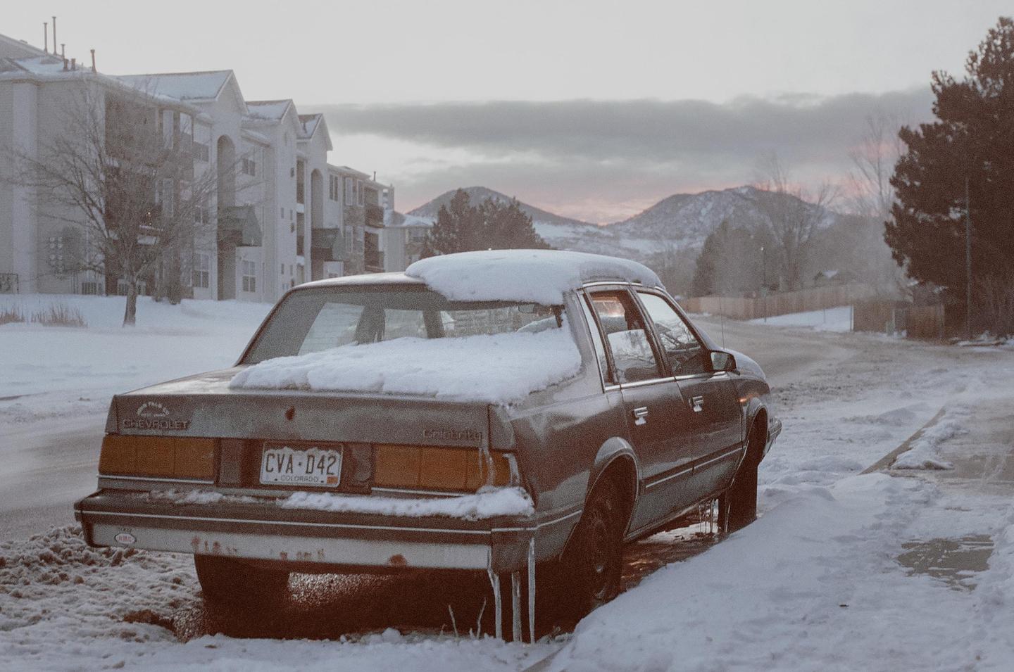 A snow-covered Chevrolet Cavalier is parked on a snowy street with mountains and buildings in the background.