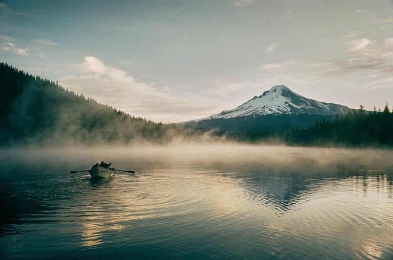 A small boat with two people rows across a misty lake towards a snow-capped mountain.