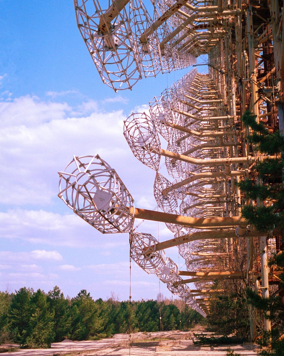 A massive, rusty Duga radar array stretches across the frame under a blue sky with scattered clouds, with a forest visible in the background.