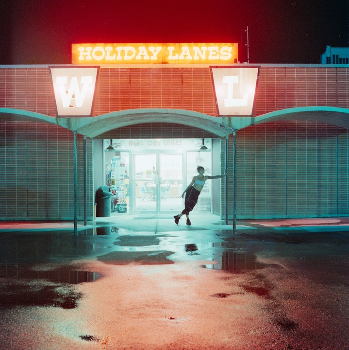 A woman stands in front of a bowling alley entrance at night, with neon signs and reflections on the wet ground.