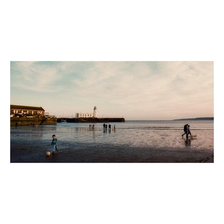A child plays with a soccer ball on a wet beach with a lighthouse and pier in the background.