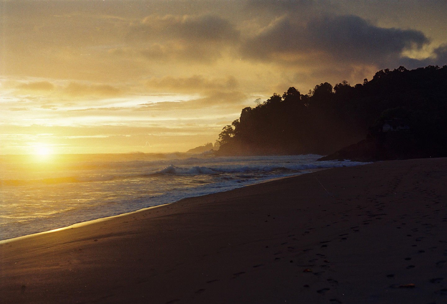 The sun sets over a beach with waves crashing on the shore and a dark, tree-covered hill in the background.