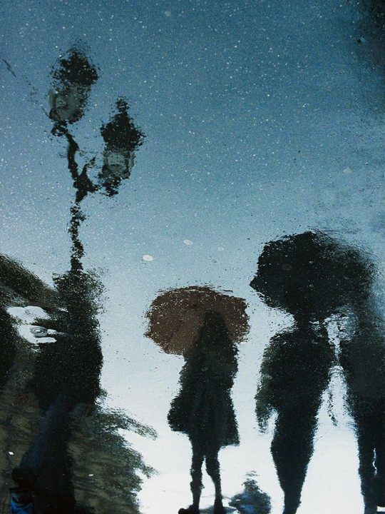 The reflection of a street light and two people, one holding an umbrella, are visible in a puddle on a street in Paris.