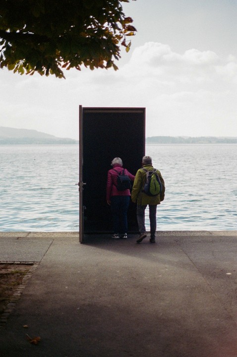 Two people stand before an open door that leads to a body of water under a cloudy sky.
