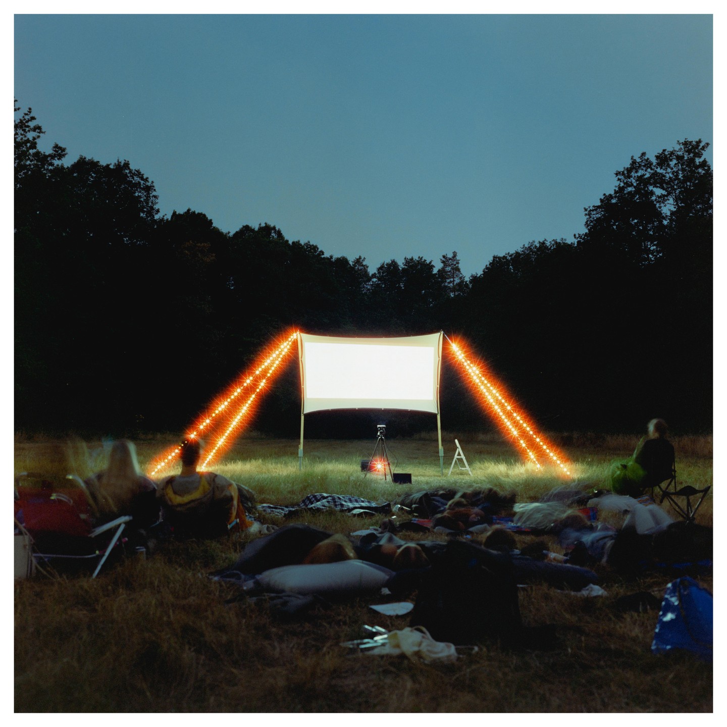 People are gathered in a grassy field at night, watching a movie projected onto a screen illuminated by string lights.