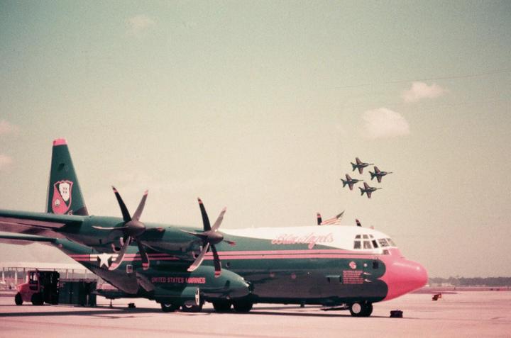 A large green and pink airplane with propellers is parked on an airfield while four smaller jets fly in formation in the sky.