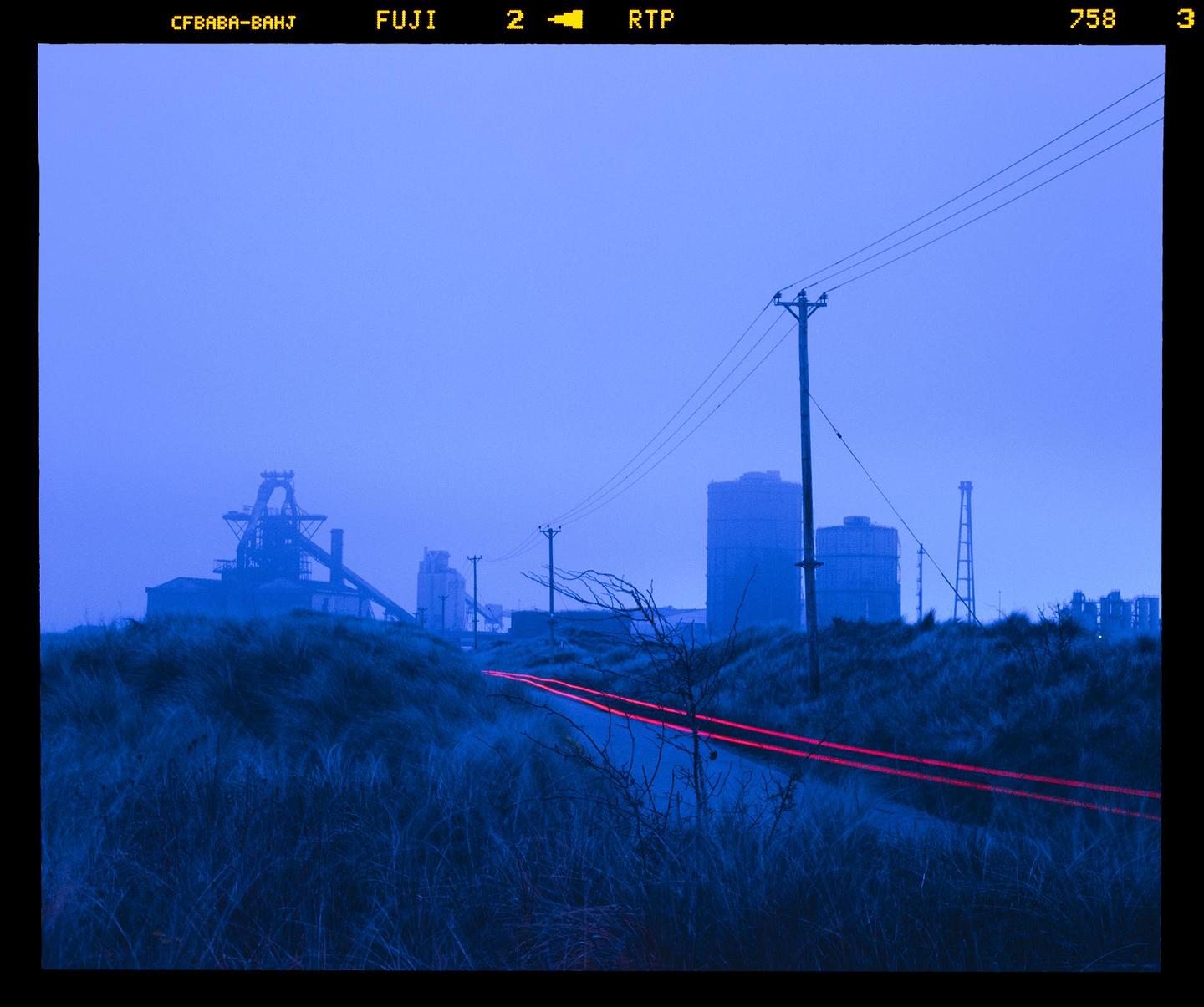 Red light trails from a vehicle on a road at dusk with industrial buildings in the background.