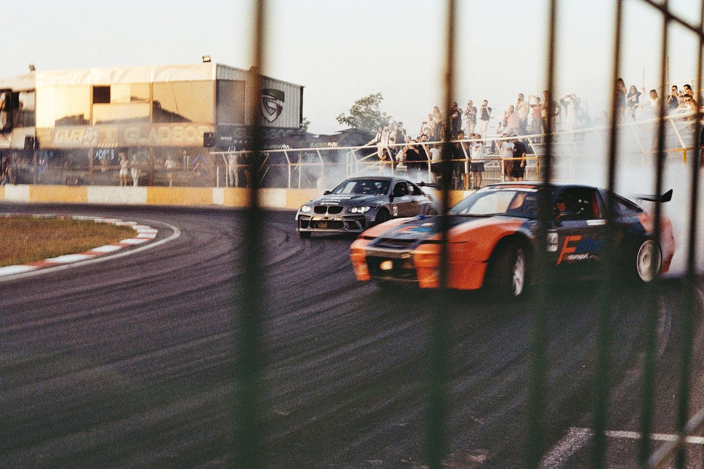 Two race cars drift around a track with spectators watching from behind a fence.