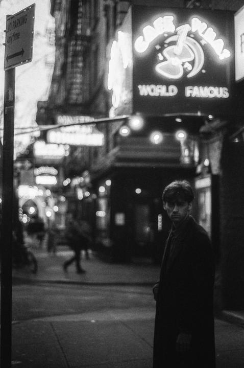 A man stands on a city street at night with neon signs and blurred figures in the background.