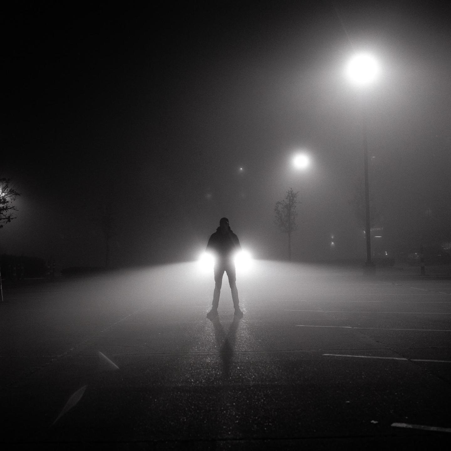 A man stands in front of a car with its headlights on in a foggy parking lot at night.