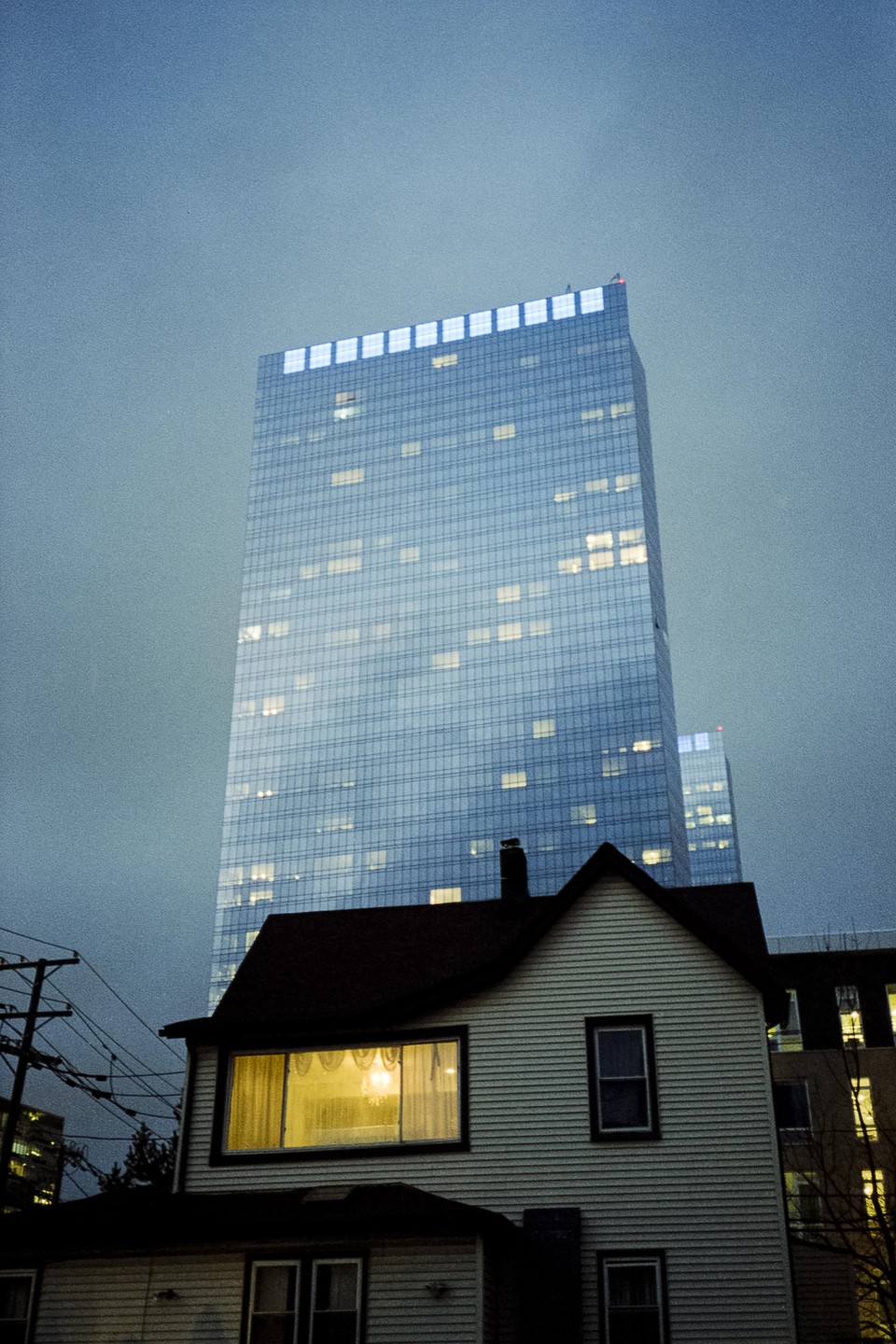 A small house with a lit window stands in front of a tall, modern skyscraper at dusk.