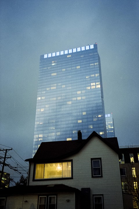 A small house with a lit window stands in front of a tall, modern skyscraper at dusk.