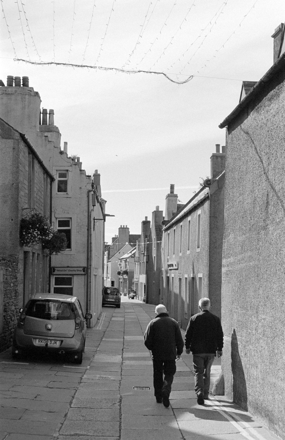 Two men walk away from the viewer down a narrow street lined with buildings and parked cars.