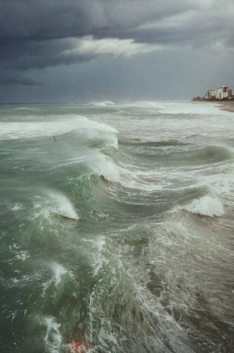 Large waves crash on a stormy ocean with buildings visible on the distant shore.