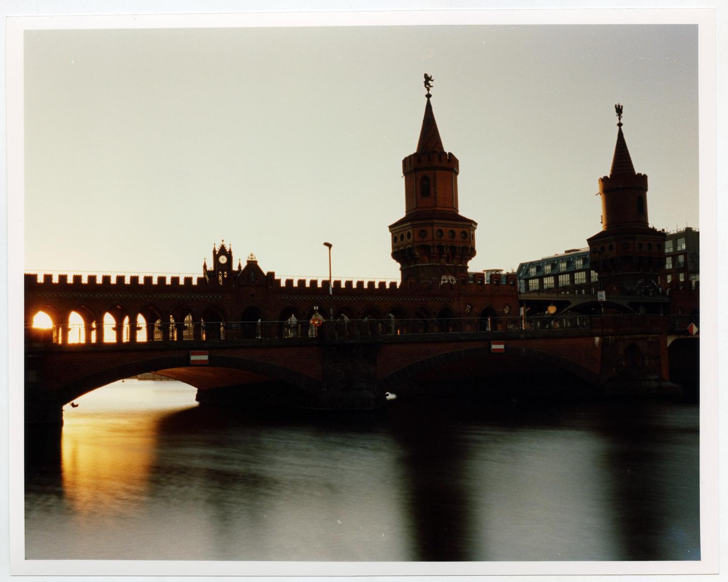 A bridge with two towers and arches is silhouetted against a bright sky, with sunlight reflecting on the water below.