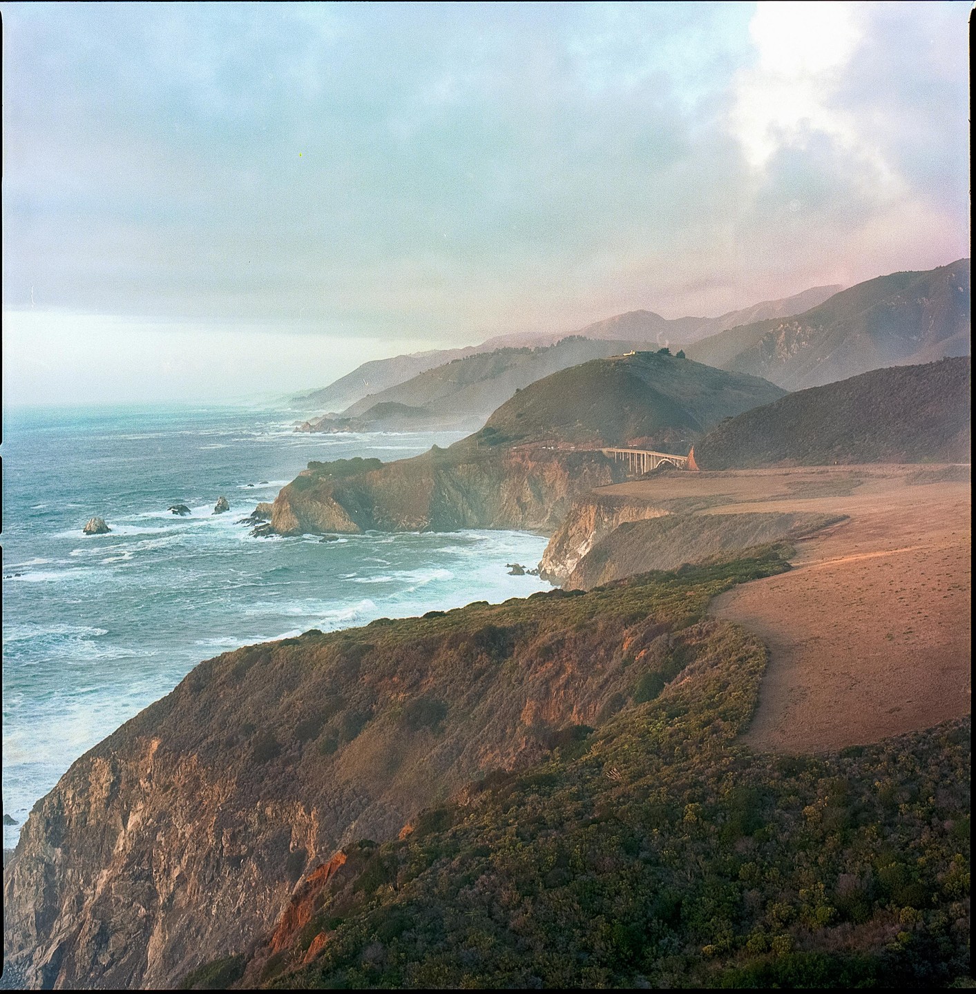 A coastal landscape features a bridge connecting two cliffs over the ocean, with mountains in the background under a cloudy sky.