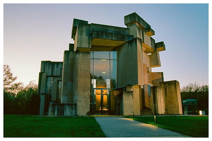 A brutalist church with geometric concrete forms stands against a twilight sky, with illuminated pathways leading to its entrance.