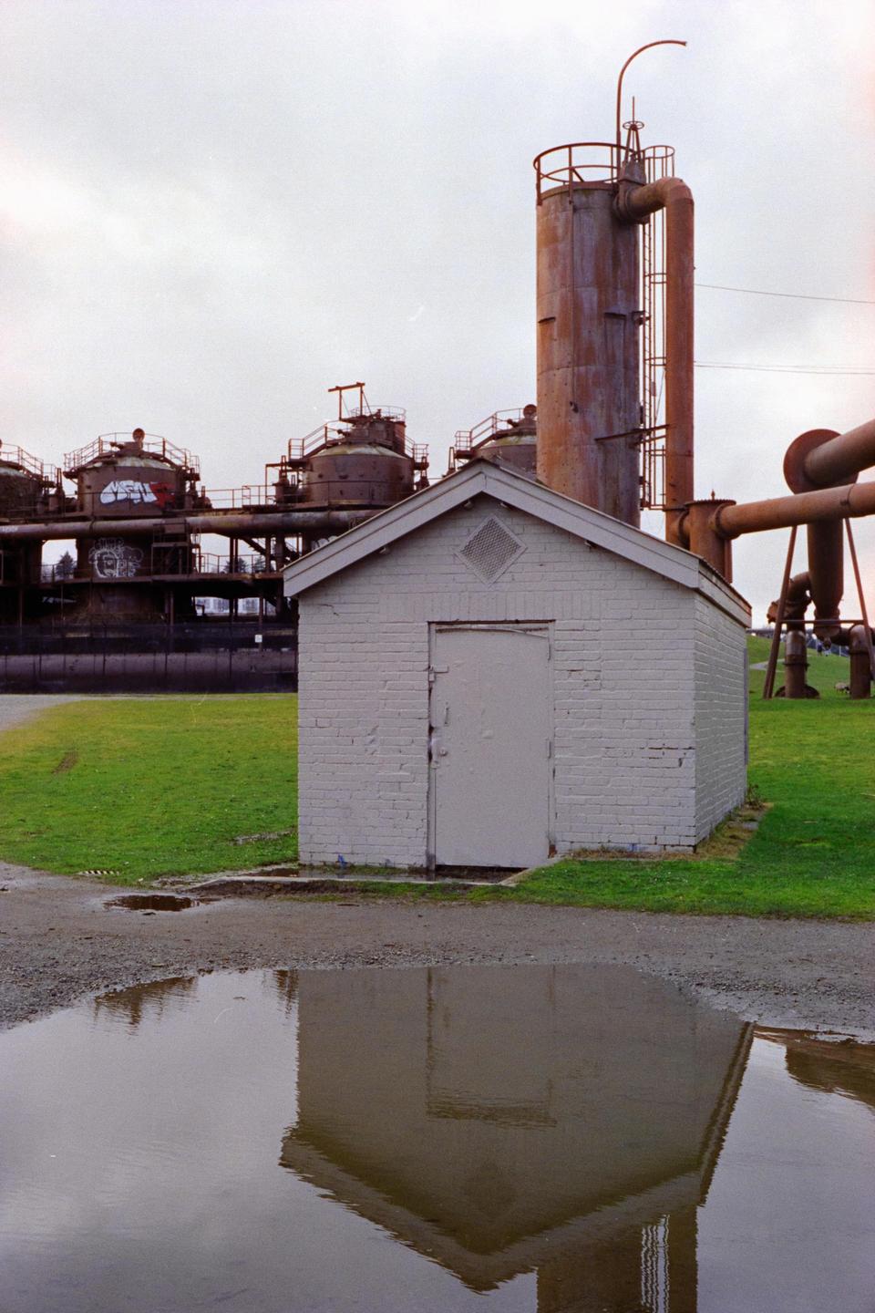 A small white brick shed stands in a grassy field with industrial structures and a puddle reflecting the shed in the background.