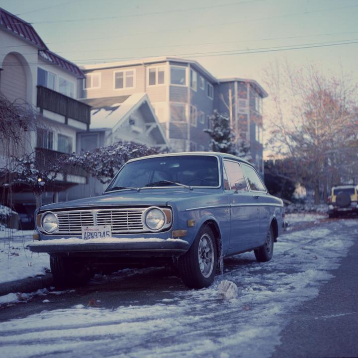 A blue car is parked on a snowy street in front of houses.