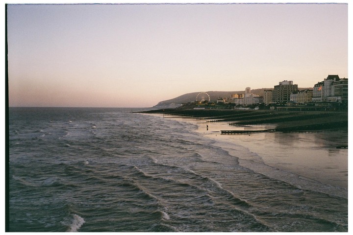 A wide shot shows a beach with waves rolling in, a person standing in the water, and a town with buildings and a ferris wheel in the background under a sunset sky.
