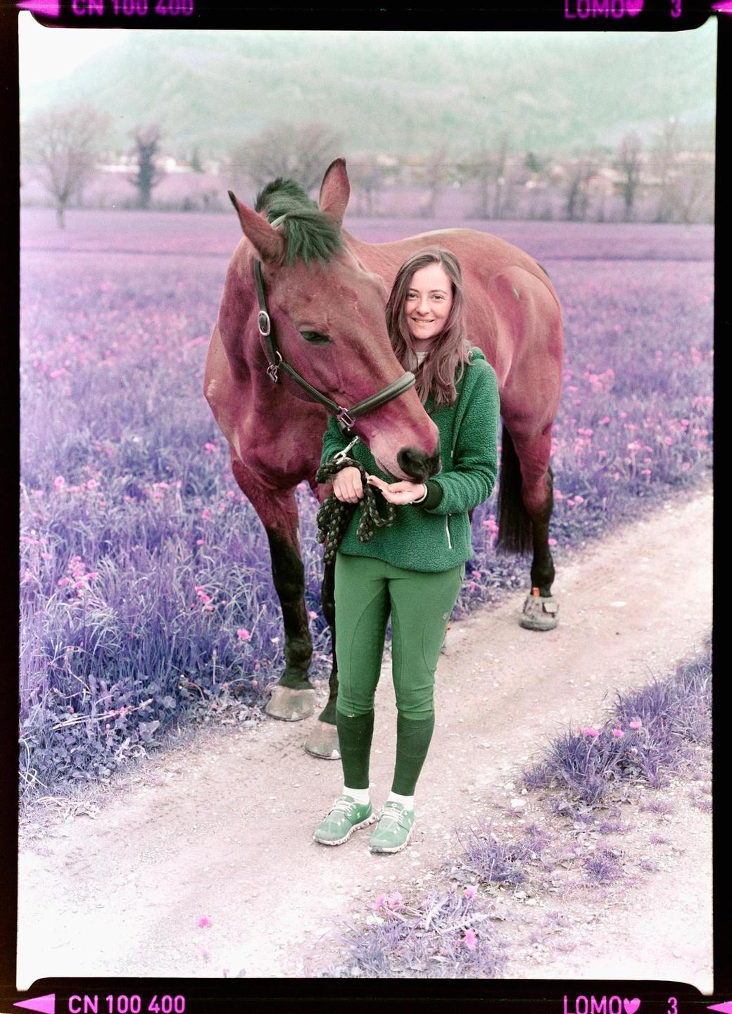 A woman stands on a dirt path next to a horse in a field of purple flowers.