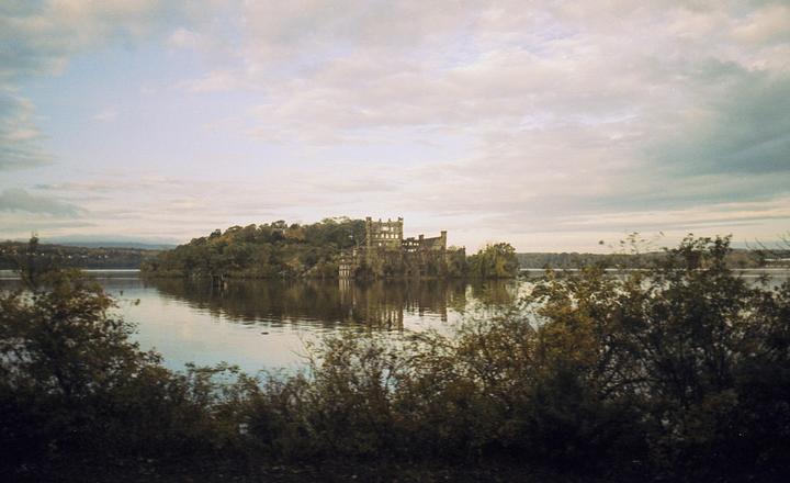 A large, ruined stone building stands on a tree-covered island in a wide river under a cloudy sky.