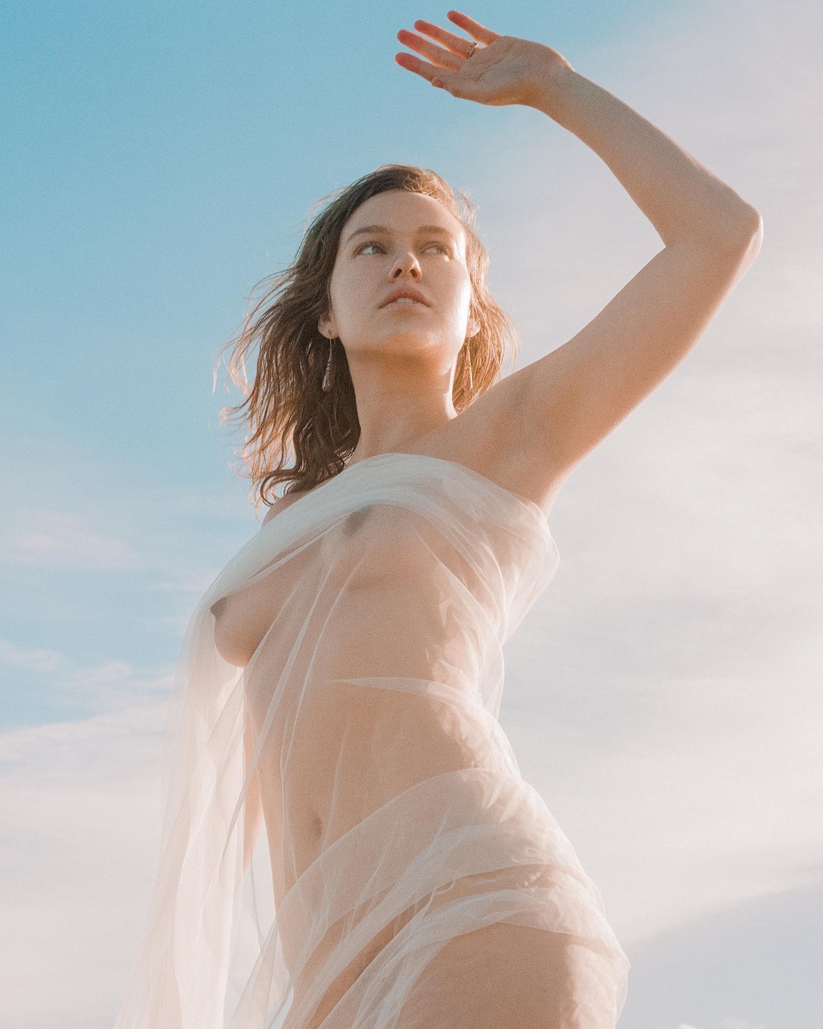 A woman with a sheer white fabric draped around her body looks up at the sky, shielding her eyes with her hand.