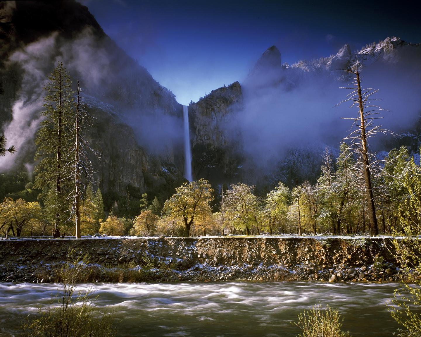 A waterfall cascades down a mountain into a river, with trees and a dusting of snow on the ground.