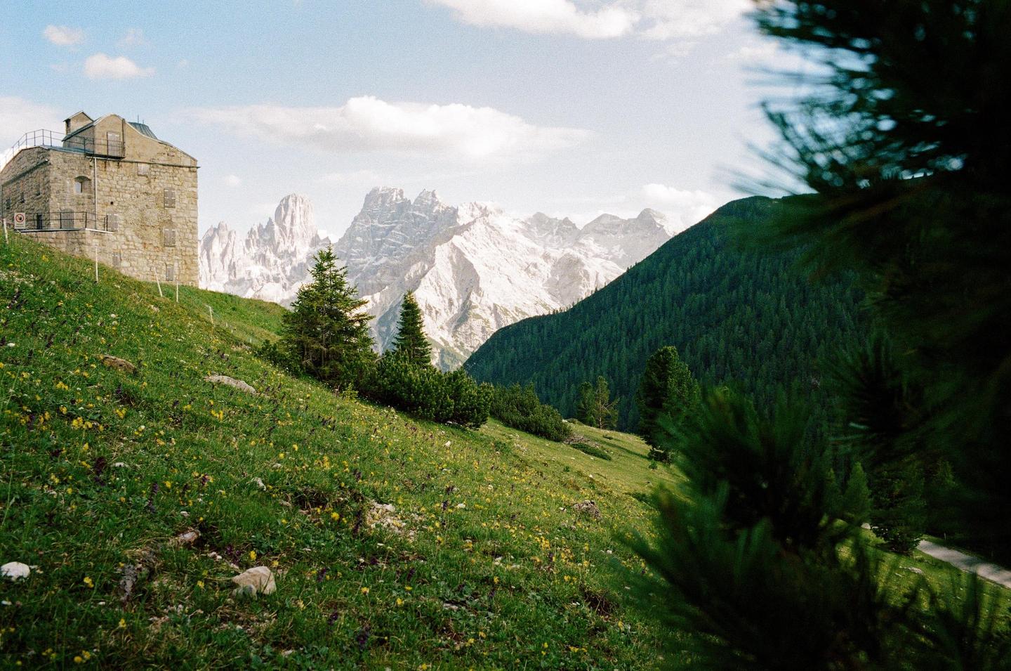A stone building stands on a grassy hillside with pine trees and mountains in the background.