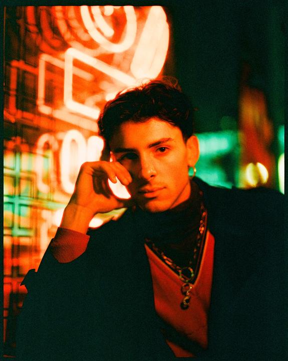 A man with dark hair and a gold earring looks at the viewer, illuminated by neon light.