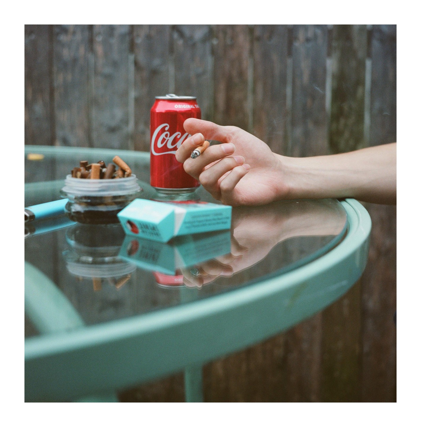 A hand holds a lit cigarette next to a can of Coca-Cola, a pack of cigarettes, and an ashtray on a glass table.