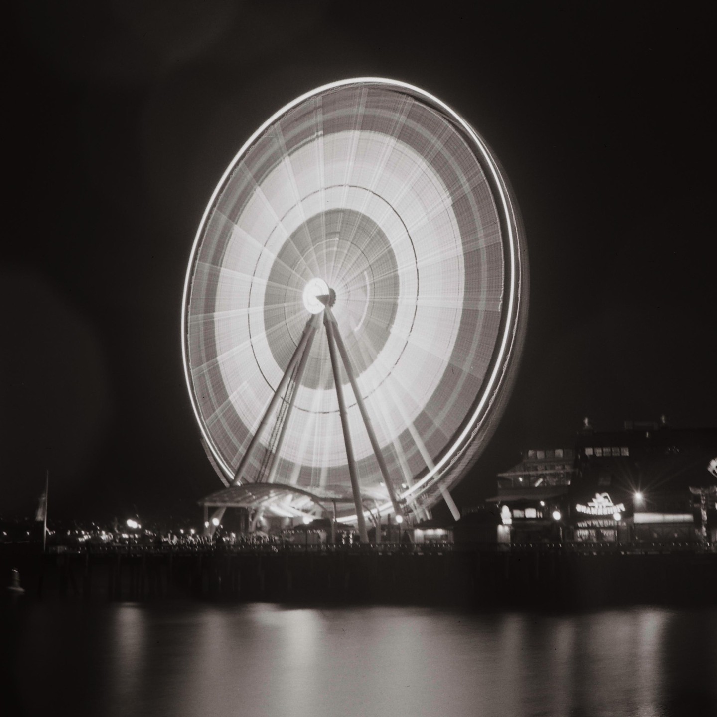 A long exposure shot of a brightly lit Ferris wheel spinning at night next to a body of water with buildings in the background.