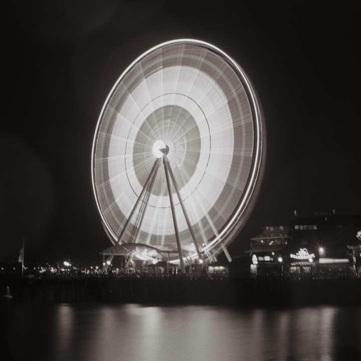 A long exposure shot of a brightly lit Ferris wheel spinning at night next to a body of water with buildings in the background.