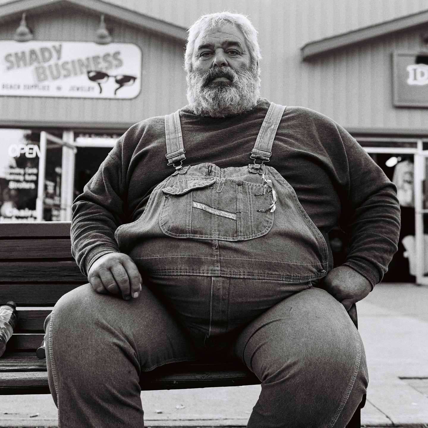 A man with a beard and overalls sits on a bench in front of a store.