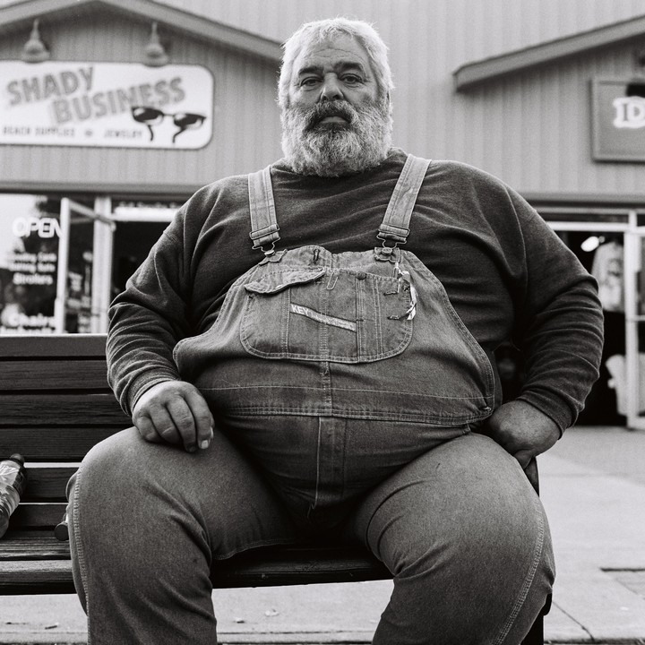 A man with a beard and overalls sits on a bench in front of a store.