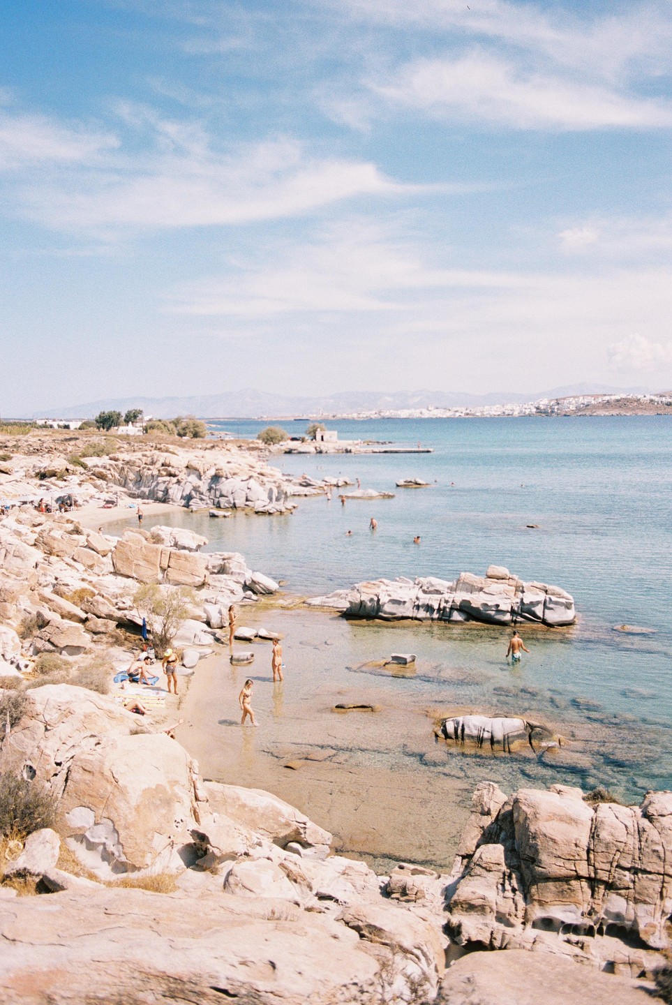 People swim and sunbathe on a rocky beach with clear blue water and a distant town under a bright sky.