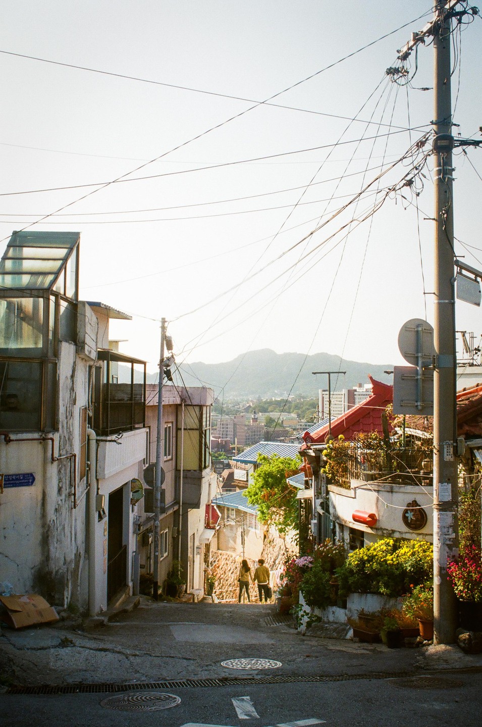A couple walks down a narrow street lined with buildings and power lines, with a city and mountains in the distance.