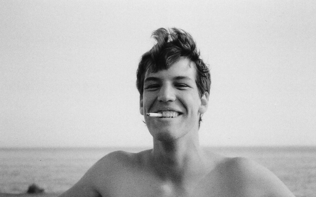 A smiling man with a cigarette in his mouth stands in front of a body of water.