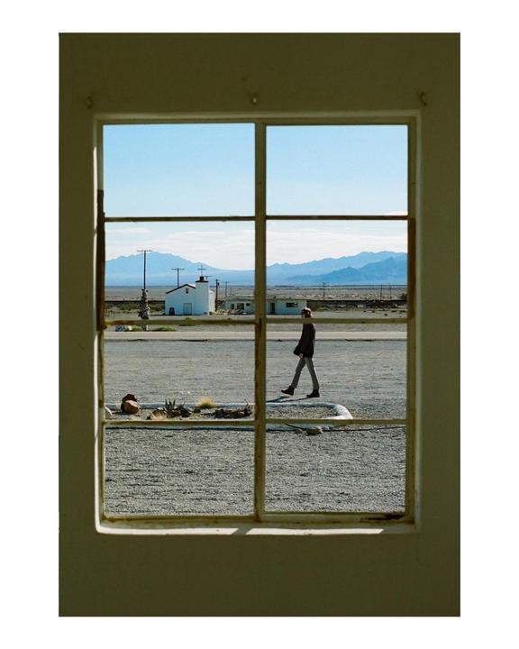 A man walks across a gravel lot towards a small white building with mountains in the background, viewed through a window frame.